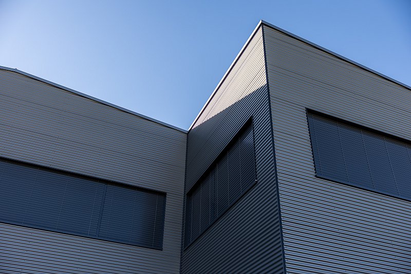 Untersicht einer kantigen Metallfassade mit dunklen Fenstern vor blauem Himmel.