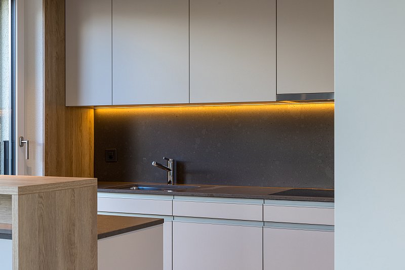 Helle Küchenschränke, Spüle und dunkle Rückwand mit warmem Lichtband.