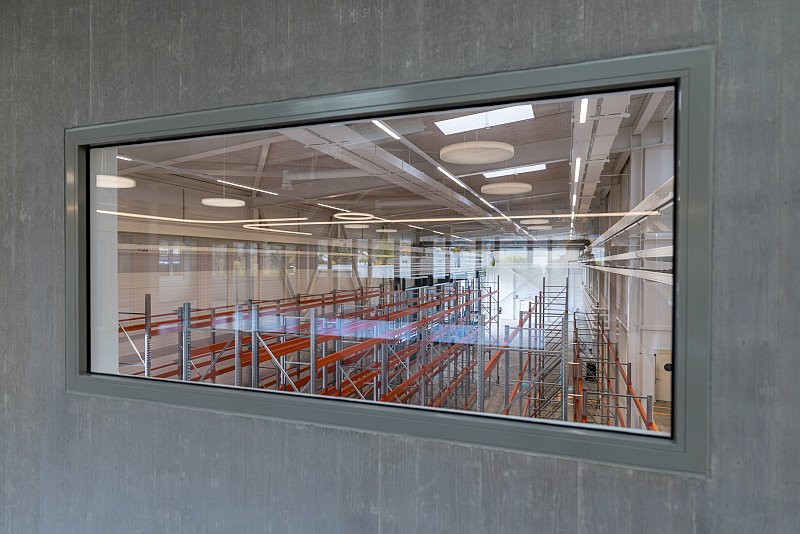 Blick durch ein Innenfenster in einer Betonwand auf eine helle Werkhalle.