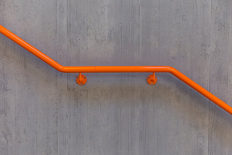 Geometrischer Verlauf eines orangefarbenen Handlaufs an einer Sichtbetonwand.