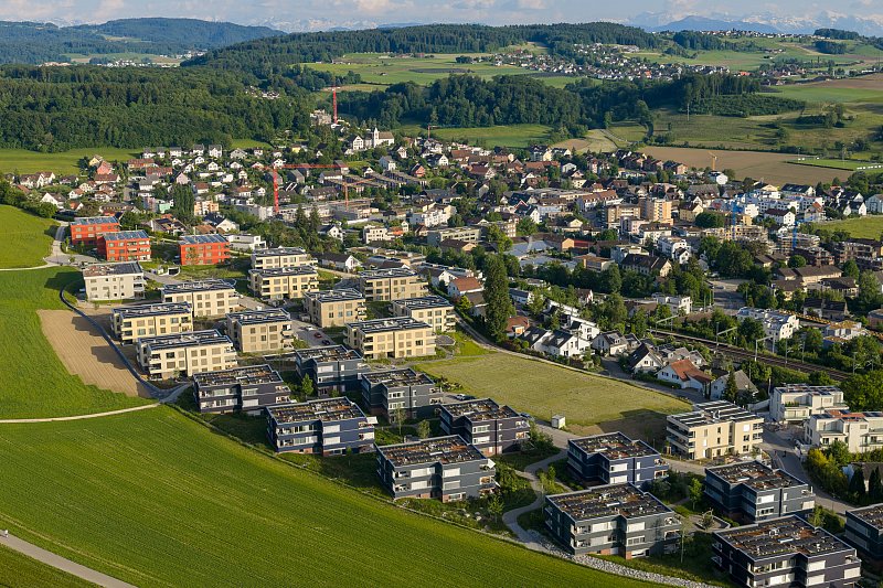 Luftaufnahme einer Wohnüberbauung am Ortsrand mit Feldern und bewaldeten Hügeln.