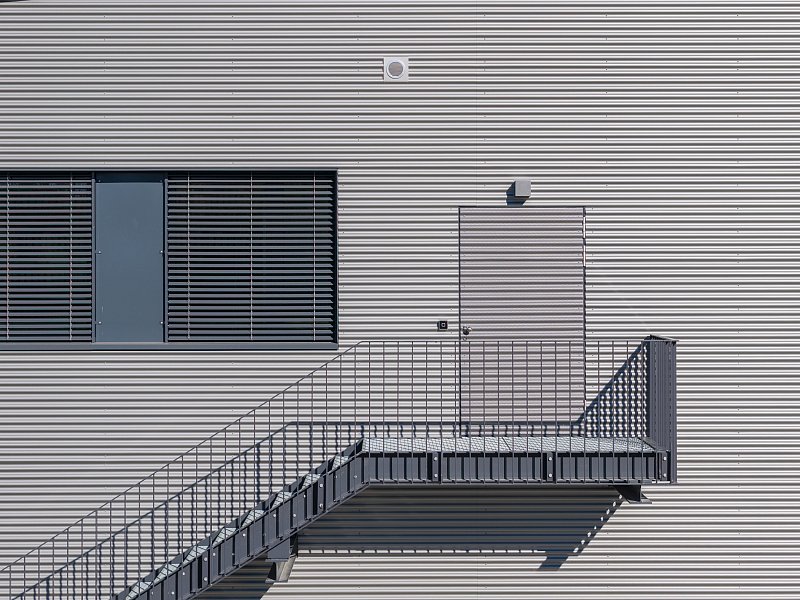 Aussentreppe mit Geländer und Schattenwurf an einer gerippten Metallfassade.