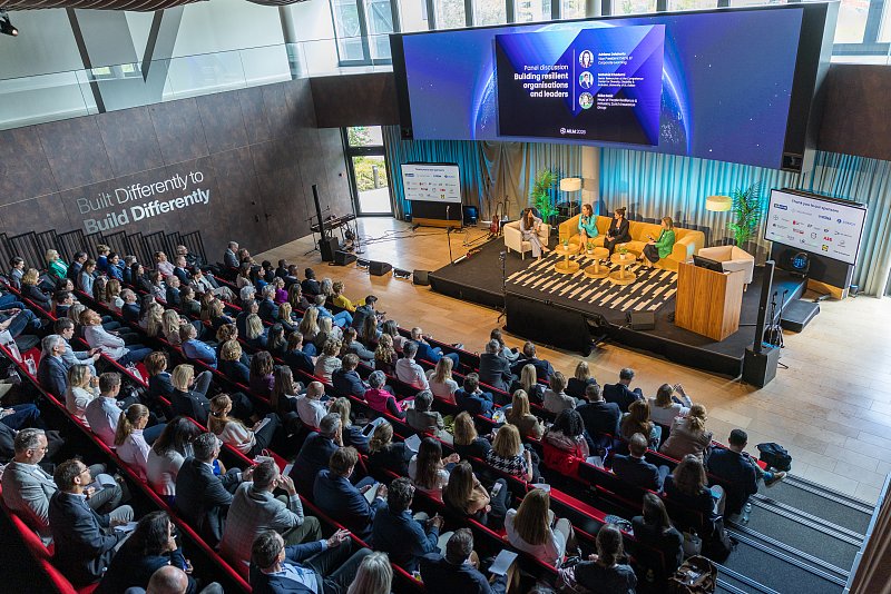 Blick von oben auf einen gefüllten Konferenzsaal mit Bühne und grossem Bildschirm.