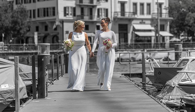 Zwei Frauen in Hochzeitskleidern gehen mit Blumen über einen Steg am Wasser.