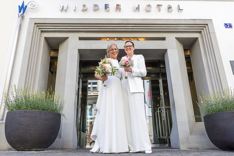 Zwei Frauen in weissen Hochzeitskleidern mit Blumensträussen vor einem Hoteleingang.