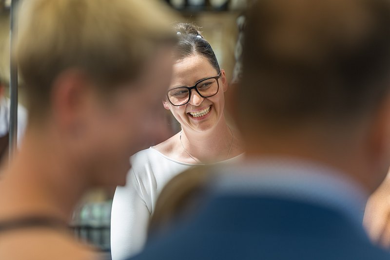Lächelnde Frau mit Brille bei einer Hochzeitsfeier, eingerahmt von unscharfen Gästen.