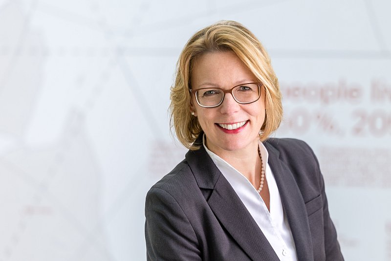 Businessporträt einer lächelnden Frau mit Brille vor einer hellen Wand.