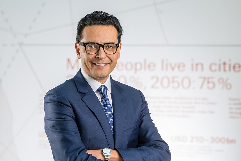Mann mit Brille im blauen Anzug vor einer hellen Wand mit Schrift.