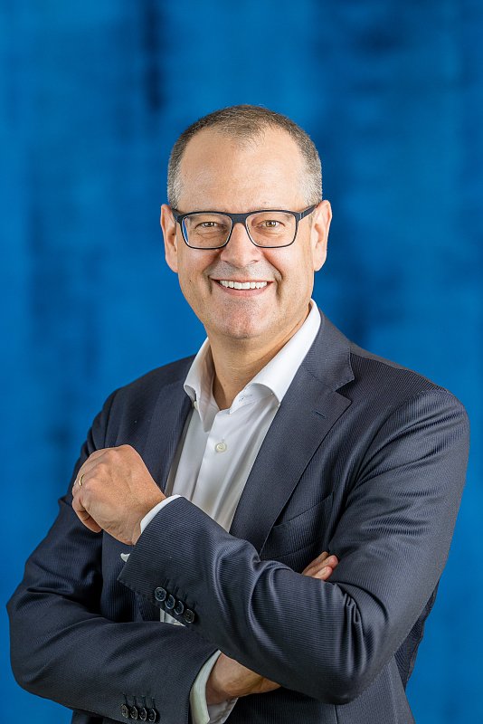 Lächelnder Mann mit Brille und verschränkten Armen vor blauem Hintergrund.