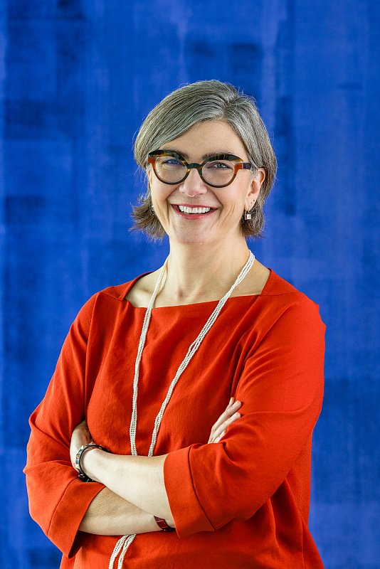 Lächelnde Frau mit Brille und roter Bluse vor blauem Hintergrund.
