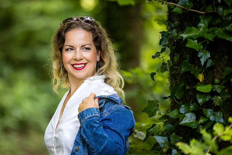 Lächelnde Frau mit Jeansjacke und weisser Bluse vor grünem Laub.