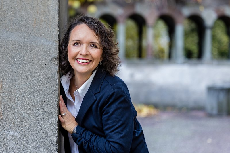 Lächelnde Frau im dunklen Blazer an einer Säule mit unscharfen Arkaden im Hintergrund.