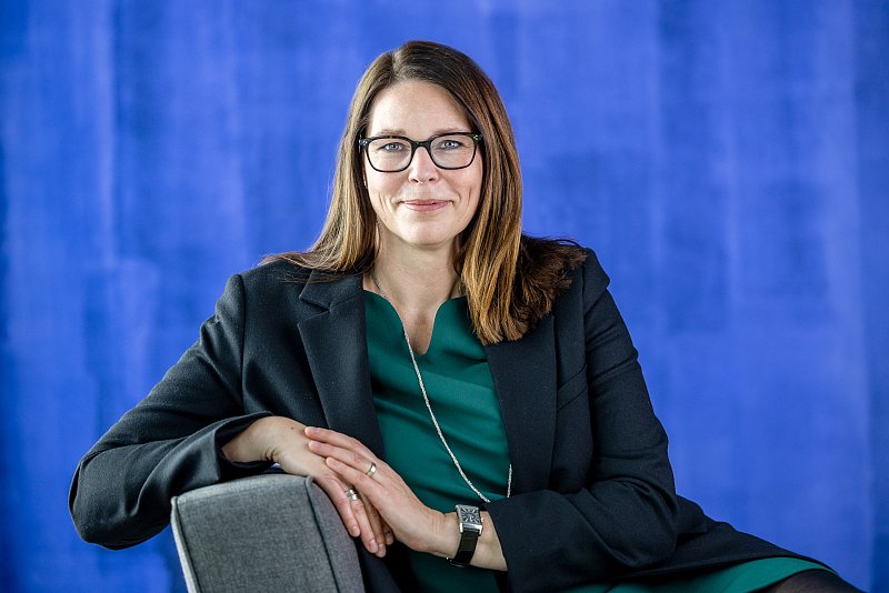 Sitzende Frau mit Brille, dunklem Blazer und grünem Oberteil vor blauem Hintergrund.