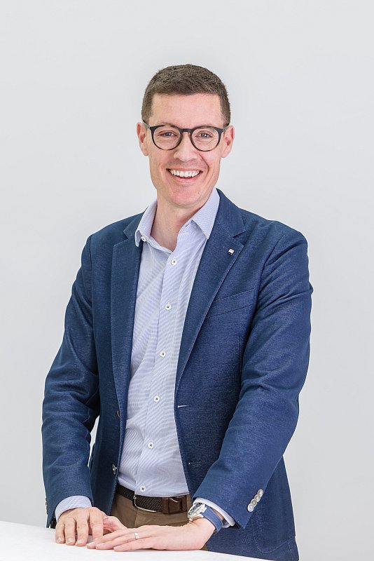Lächelnder Mann mit Brille und blauem Blazer stützt sich auf einen hellen Tisch.